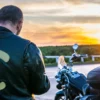 Rider in a leather motorcycle jacket with reflective panels beside his bike at sunset